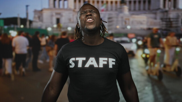 Young african american man wearing staff shirt with open mouth shouting and hands raised, dreadlocks visible in crowded city street; passion urban energy.