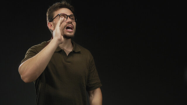 Young hispanic man wearing glasses and brown polo with raised hand shouting and cupping mouth in black studio; urgent plea urgency.