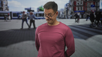 Fototapeta premium Man with glasses and moustache smirks, tilts his head and keeps hands behind his back on a busy street crosswalk among trams and cyclists; playful mischief.