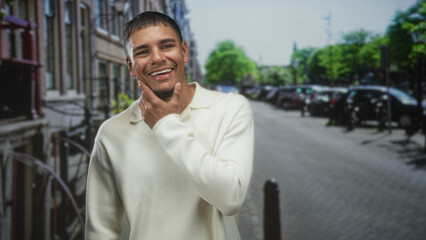 Fototapeta premium Young hispanic man with hand on chin smiling and covering mouth in a soft laugh, wearing cream sweater on street; shy amusement.