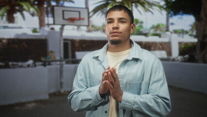 Obraz premium Man clasping hands at chest in light denim shirt beside a basketball hoop on a street court, young hispanic male with focused gaze and trimmed goatee; quiet determination.