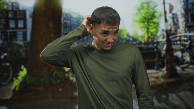 Man wearing green long sleeve shirt scratching head with hand and wry smile on cobbled street by canal and bikes, face and hand visible; awkward charm.