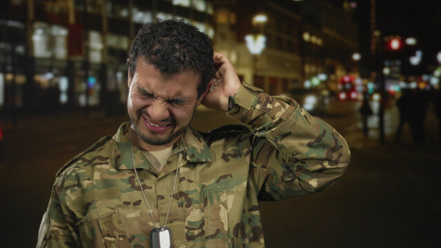 Man wearing camouflage uniform and dogtags grimaces on busy city street at night under glowing streetlights; weariness.
