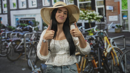Fototapeta premium Young hispanic woman wearing straw hat and white blouse shows both thumbs up and open palms beside a crowded bicycle rack on a street; travel joy.
