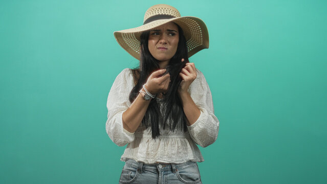 Young hispanic woman in straw sunhat holding long hair with both hands and grimacing while inspecting split ends in studio; disgust.