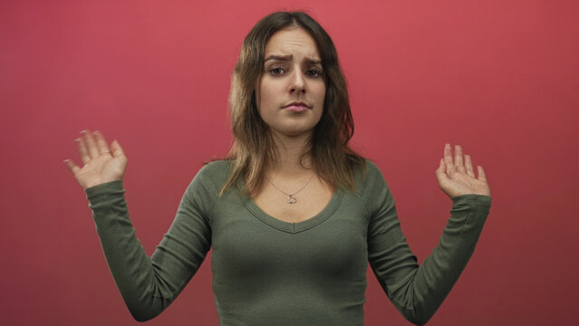 Young hispanic woman with heart necklace raises both hands in a shrug gesture in a pink studio; resignation.