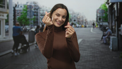 Fototapeta premium Woman smiling and making finger heart gesture with hands near chest in a brown turtleneck on a busy cobblestone street in city center; joy warmth.