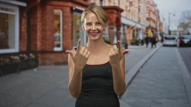 Woman smiling with hands showing call me gesture, thumbs and pinkies extended, eyes closed, wearing black tanktop in street with brick building facades and sidewalk; playful invitation.