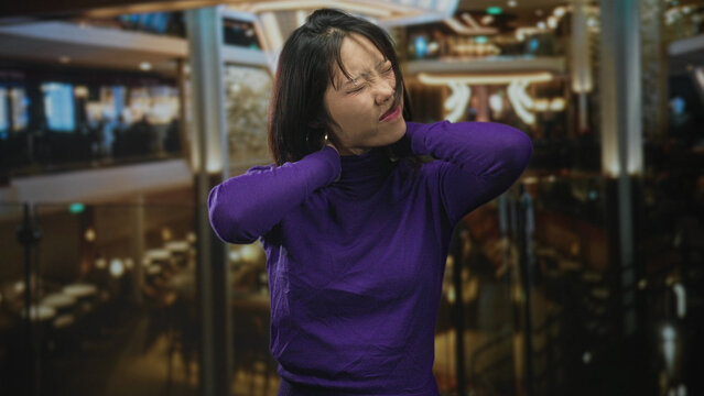 Woman holding neck with both hands in building, wearing purple turtleneck and earrings while wincing; discomfort tension.