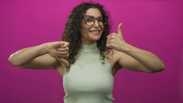Young hispanic woman smiling with eyes closed showing thumbs up and thumbs down in pink studio; ambivalence.