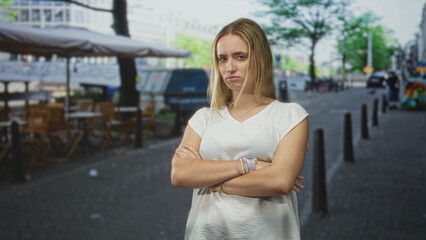 Fototapeta premium Woman with arms crossed and visible wristwatch, frowning on a street cafe scene; disapproval resolve.