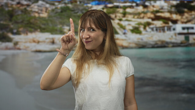 Woman pointing index finger toward camera in studio setting with seaside backdrop and white t shirt visible; playful confidence.