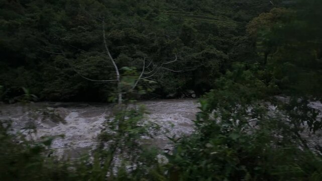 View from a train traveling toward Aguas Calientes, Peru, on a rainy and overcast day, with a powerful river rushing alongside the tracks through the dramatic mountain landscape