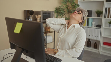 Woman in white shirt wearing glasses holding neck leaning back at office desk computer monitor; fatigue.