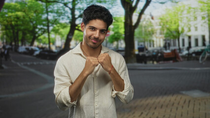Fototapeta premium Man with fists raised near chin revealing bare forearms on tree lined street, wearing linen shirt and red bracelet; confidence friendly.