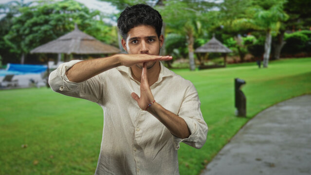 Man showing a timeout hand gesture with palms and forearms visible in a forest setting beside a paved path and thatched huts; determined refusal.