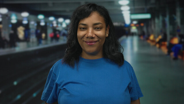 Woman in blue shirt standing on platform at train station under overhead lights smiling gently; quiet confidence.