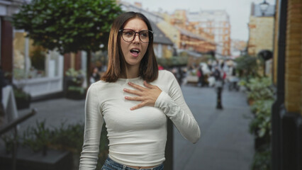 Fototapeta premium Young hispanic woman hand on chest gesture on a busy pedestrian street with shops and planters; surprise vulnerability.