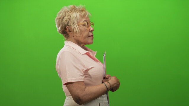 Senior woman in pink polo holds clipboard with hands clasped in profile at studio green screen; calm focus.