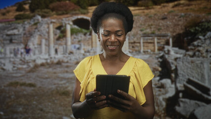 Fototapeta premium Woman holds tablet with both hands, tapping screen while standing among ancient ruins building and reading; curiosity travel discovery.