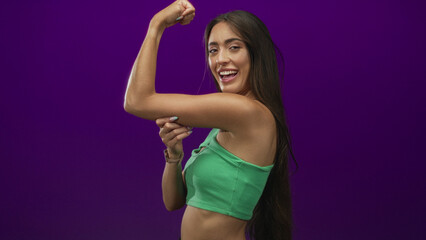 Obraz premium Young hispanic woman flexing biceps in purple studio wearing green crop top and smiling; confidence empowerment fitness.