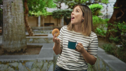 Fototapeta premium Young woman holding a glazed donut and teal enamel mug with mouth open in a green forest courtyard near stone wall; surprise breakfast.