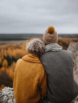 A loving couple in cozy sweaters sits at a countryside house, gazing at vibrant autumn landscape with yellow and red leaves. Calm lifestyle, togetherness with nature and each other. View from behind.