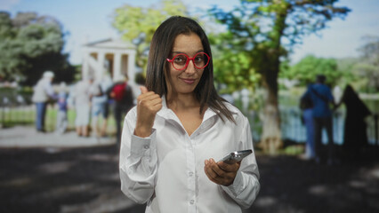 Fototapeta premium Woman holding smartphone and pointing thumb at camera in forest setting while wearing red glasses and white shirt; confidence connection ease.