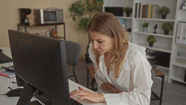 Woman typing on keyboard at a desk in a building while clutching her abdomen for stomachache and pausing between keystrokes; discomfort.