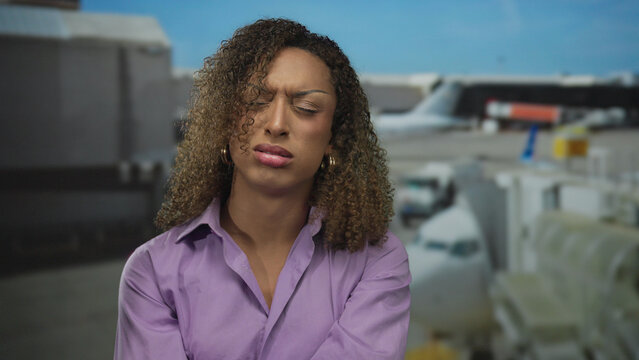 Woman with curly hair crosses arms at airport terminal outdoors under blue sky near boarding gate; rejection.