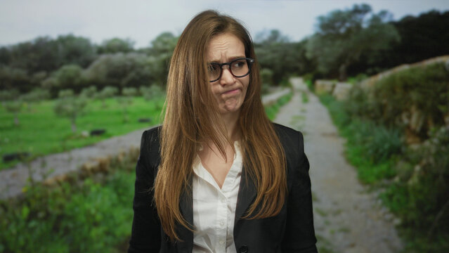Young woman furrows brow with downcast eyes and pouty lips in black blazer and white shirt while standing on a leafy park trail; sadness.