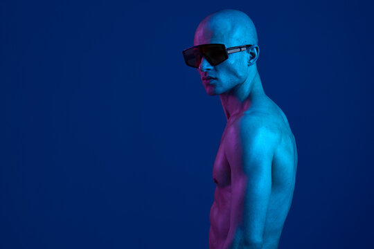 Bald man posing confidently in blue lighting with stylish sunglasses, showcasing a strong physique in a vibrant atmosphere. Perfect for fashion and advertising needs.