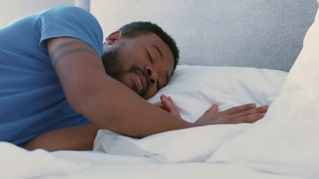 A man with dark skin is sleeping peacefully on a bed. Morning light reflects softly on him as he relaxes. The comfortable setting creates a sense of rest and comfort.