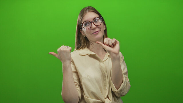 Smiling woman pointing both thumbs at her chest in green studio setting with eyeglasses and beige blouse; pride.