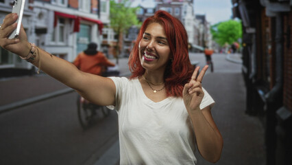 Fototapeta premium Young woman smiling taking selfie with smartphone and peace sign in white t shirt on narrow city street; joy social sharing.