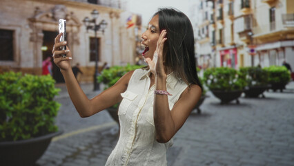 Fototapeta premium Thai woman wearing a sleeveless white top holds smartphone and flashes a peace sign while smiling on a cobblestone street; joyful travel moment.