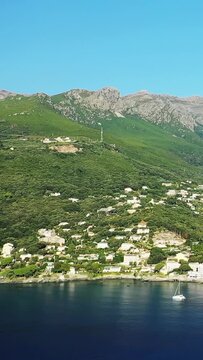 Scenic view of a coastal village nestled between lush green hills and mountains, with a sailboat on calm blue water in Erbalunga, Corsica.
