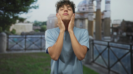 Fototapeta premium Young man rubbing hands over face near stone building, closed eyes and relaxed posture; quiet contemplation.