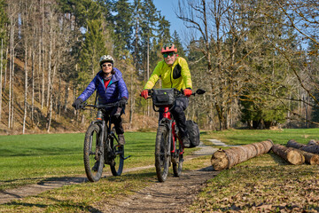 Naklejka premium Happy senior couple enjoying an active lifestyle, riding electric bikes on a forest trail in spring. Outdoor recreation, healthy aging, and leisure time in nature on a sunny day.