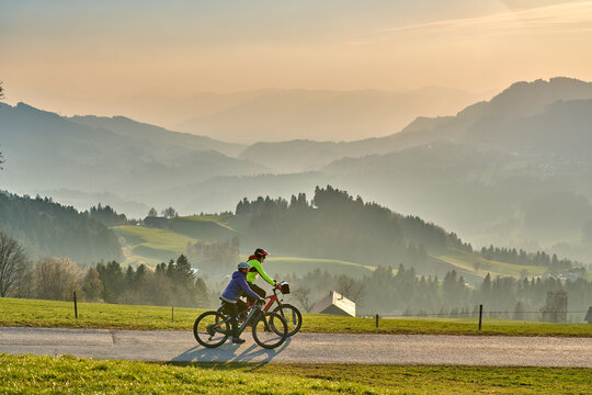 Active senior couple riding electric bikes through scenic Bregenzerwald in Austria during golden evening light. Enjoying nature, mountains, and healthy outdoor lifestyle in spring countryside.
