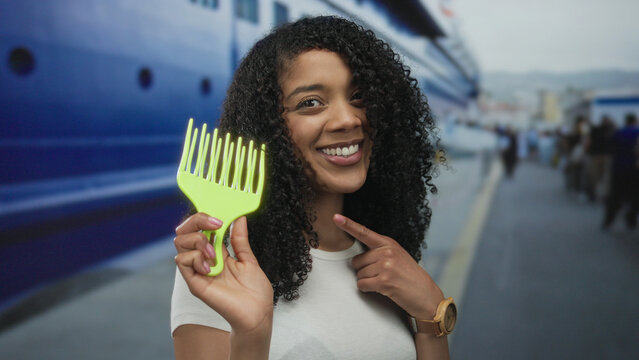 African american woman with curly hair holds bright green pick comb and points finger to comb on street; confidence.