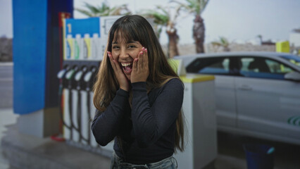 Woman smiling with hands raised at gas pump on street wearing dark sweater and visible necklace, car and fuel dispensers behind her  joy. © Krakenimages.com