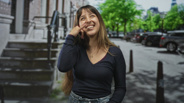 Young woman pointing with hand and miming phone gesture on street, wearing necklace and dark vneck, long hair, smiling sideways; playful smile joy.