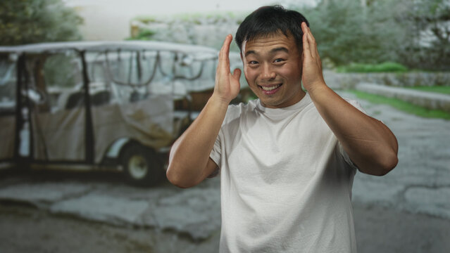Smiling man covers ears with hands on a cracked street beside a parked tuk tuk outdoors; playfulness.