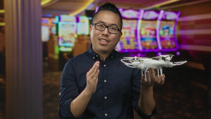 Fototapeta premium Man holding white quadcopter drone in one hand and gesturing with other while speaking in casino building with slot machines visible; curiosity.