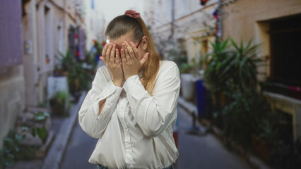 Fototapeta premium Woman covering hands over face on a narrow urban street lined with potted plants and doorways; embarrassment vulnerability.