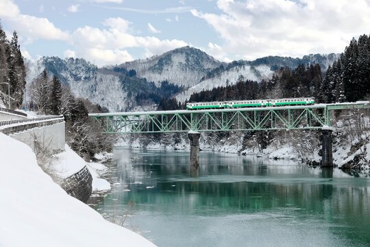 A local train of JR Tadami railway line traveling on a bridge over Tadami River after heavy snowfalls on a cold, gloomy winter morning in Kaneyama Machi, Oku-Aizu, Fukushima, Tohoku, Japan