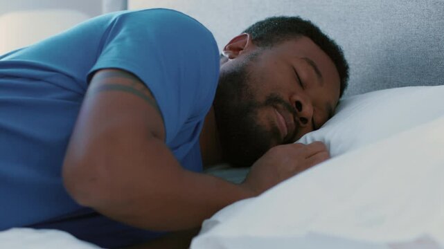 A man with dark skin is lying on his side, sleeping comfortably in a well-lit bedroom. The setting shows soft bedding and a cozy atmosphere, suggesting rest and relaxation.
