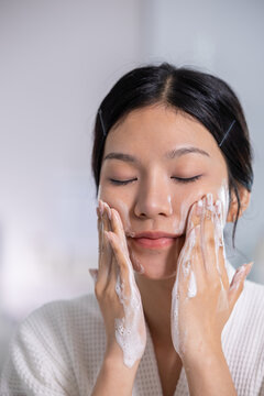 Beautiful young Asian woman washing face with cleansing foam enjoying morning self care routine feeling relaxed and peaceful with closed eyes wearing white robe