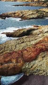Waves crash against rugged rocky cliffs along the coastline at Delta du Fango in Corsica, France. Natural seascape with textured rocks and turquoise water.